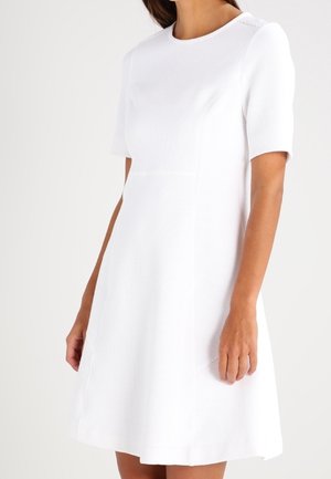 Kobieta ubrana w białą, teksturowaną sukienkę o krótkim rękawie sięgającą kolan, z okrągłym dekoltem, stojąca na tle jasnej, jednolitej ściany.