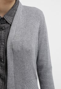 Cardigan en maille côtelée grise porté sur une chemise noire à boutons pression, montré de la poitrine à l'épaule sur une personne.