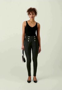 Black sleeveless top paired with high-waisted dark trousers featuring two large silver buttons. Model holds a small black handbag, wearing black heels.