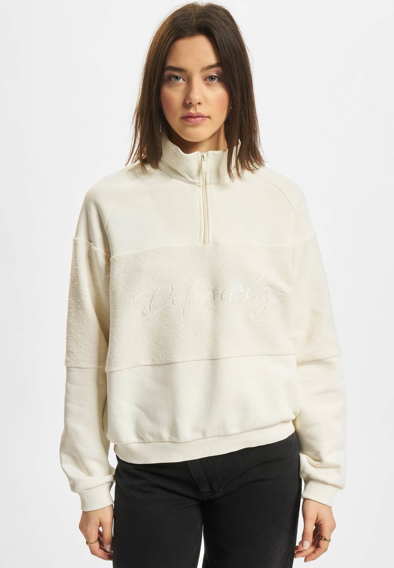 Weißes Sweatshirt mit halbem Reißverschluss, aus texturiertem Stoff über der Brust und einem gestickten Akzent mit der Aufschrift "Definitiv." Lässiger Schnitt.