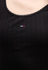 Czarny prążkowany top na ramiączkach z falbaniastym dekoltem, z małym haftowanym logo w czerwono-białoniebieskich kolorach na piersi.