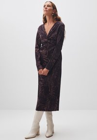 Langes, lila-schwarz gestreiftes Kleid mit tiefem V-Ausschnitt und Twists detail an der Taille, kombiniert mit weißen, hohen Stiefeln.