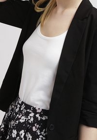 Blazer noir porté sur un débardeur blanc, associé à une jupe fleurie avec de petites fleurs roses et blanches sur un fond sombre.