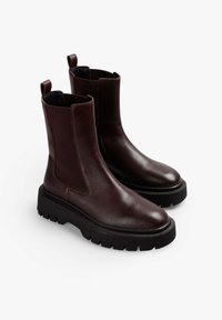 Botas Chelsea de cuero marrón con paneles laterales elásticos, tirador y suelas de goma negra gruesas. Textura suave con un diseño elegante.