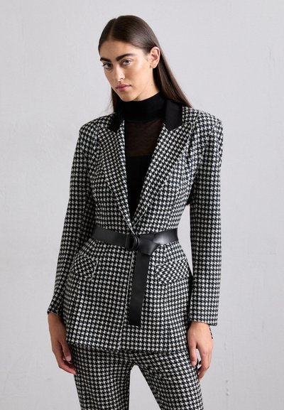 Valkoinen ja musta houndstooth-sakkihousupuku, jossa on hyvin istuva leikkaus, yksinäinen nappi kiinnitys ja musta vyö. Tyylitelty mustan poolopaidan päälle.