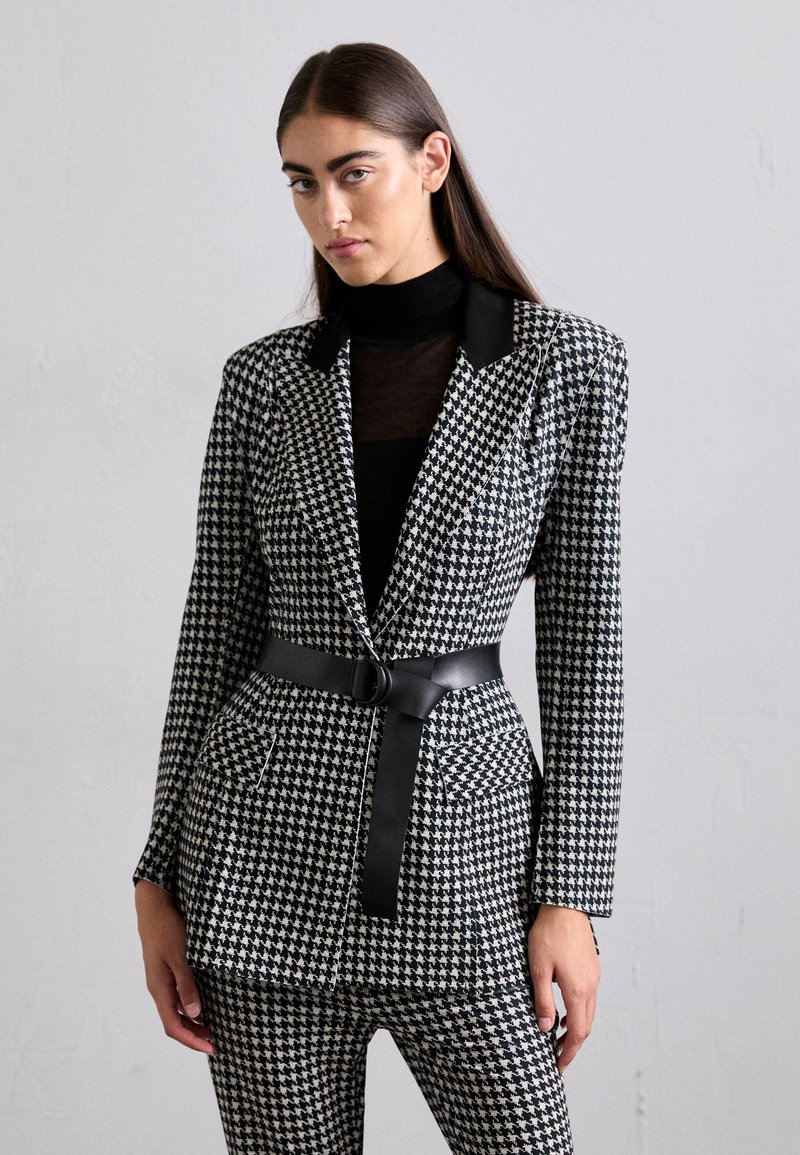 Fekete-fehér houndstooth mintás blézer, testhez simuló fazonnal, egysoros gombolással és fekete övvel. Fekete garbó fölé van stílusosan viselve.