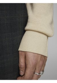 Beige gestrickter Ärmel mit geripptem Bündchen neben einem dunklen Karo-Stoff. Sichtbare Hand, die einen silbernen Ring trägt, zeigt subtile Details und Texturen.