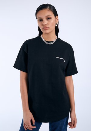 Młoda kobieta z włosami w mokrym stylu nosi czarną koszulkę z małym białym napisem, srebrny łańcuszek i kolczyki w kształcie serca.