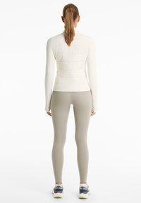 Femme debout de dos, portant une veste matelassée blanche, un legging gris clair et des baskets gris-bleu avec des semelles blanches.