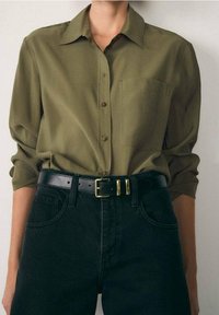 Chemise boutonnée à manches longues de couleur vert olive avec une poche, associée à un jean en denim noir taille haute et une ceinture noire avec une boucle dorée.