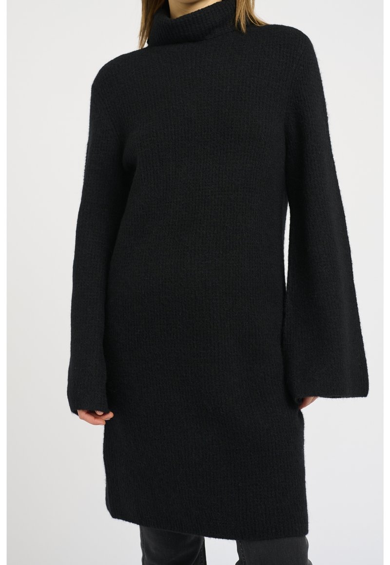 Robe pull noire en maille avec col roulé et manches longues larges portée sur un pantalon sombre sur fond clair.
