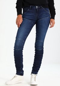 Smala jeans i mörkblå denim har en hög midja, texturerade detaljer längs benen och ribbade accenter vid knäna. Matchas med vita sneakers.