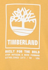 Wit Timberland-logo met gestileerde boom boven de merknaam en slogan "Gebouwd voor de moedigen, stap naar buiten en ga vooruit" op een gele achtergrond.