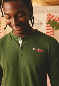 Grön långärmad skjorta med en texturerad yta, som har en flerfärgad blommig innerkant och ett broderat röd bilmönster.