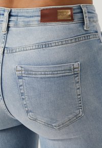 Detailní záběr na zadní kapsu světle modrých džín s viditelným šitím a hnědou značkovou nášivkou na pase.