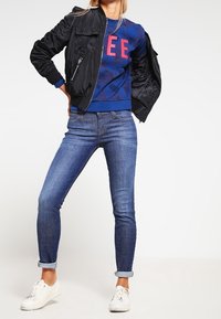 Mujer con chaqueta bomber negra, sudadera azul con letras rojas, jeans ajustados arremangados y zapatillas blancas frente a un fondo blanco.