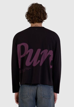 Person with curly hair facing away wearing a black long-sleeve shirt with large purple text and blue denim jeans.
