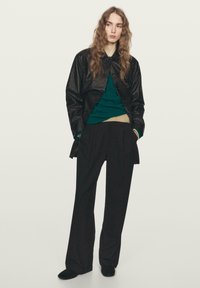 Schwarze, strukturierte Jacke über einem grünen Pullover und einem beigefarbenen Oberteil, kombiniert mit Hose mit Nadelstreifen. Schwarze Schuhe vervollständigen das Outfit.