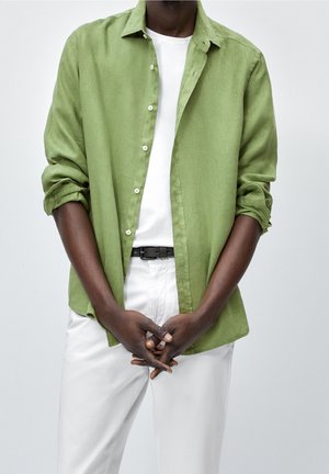 Man wearing rolled-up sleeves green button-up shirt over white T-shirt and white pants with black belt, hands clasped in front.