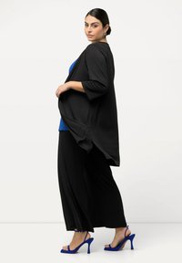 Svart öppen cardigan över en blå topp, parat med svarta luftiga byxor och blå högklackade sandaler. Slätt tyg, avslappnad passform och trekvartsärmar.