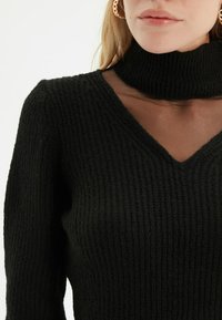 Maglione nero a coste con scollo a V e alto collo a dolce vita, caratterizzato da un dettaglio in rete trasparente all'collo.