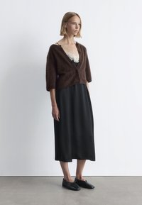 Brauner, flauschiger Strickcardigan mit Dreiviertelärmeln, getragen über einem schwarzen satinierten Slipkleid mit Spitzenakzenten und schwarzen flachen Schuhen.