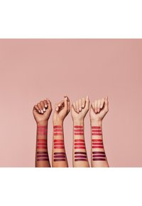 Fire armer viser leppefarger i forskjellige nyanser, inkludert rosa, rød og lilla, på en lys rosa bakgrunn. Hudtoner varierer.