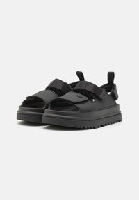 Sorte sandaler med en kraftig såle, laget av syntetisk materiale. Har justerbare stropper og utskårne sider for bedre pusteevne.