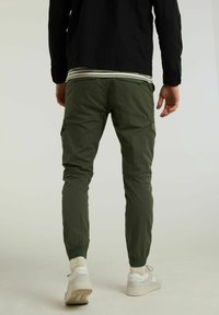 Adulto vistiendo pantalones jogger verde oliva, calcetines blancos, zapatillas blancas, chaqueta negra y camiseta a rayas, de pie contra un fondo claro y liso.