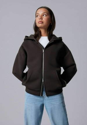 Bruine gewatteerde hoodie met een rits aan de voorkant en twee zakken, gecombineerd met lichtblauwe spijkerbroek. Glad textuur en een ontspannen pasvorm.