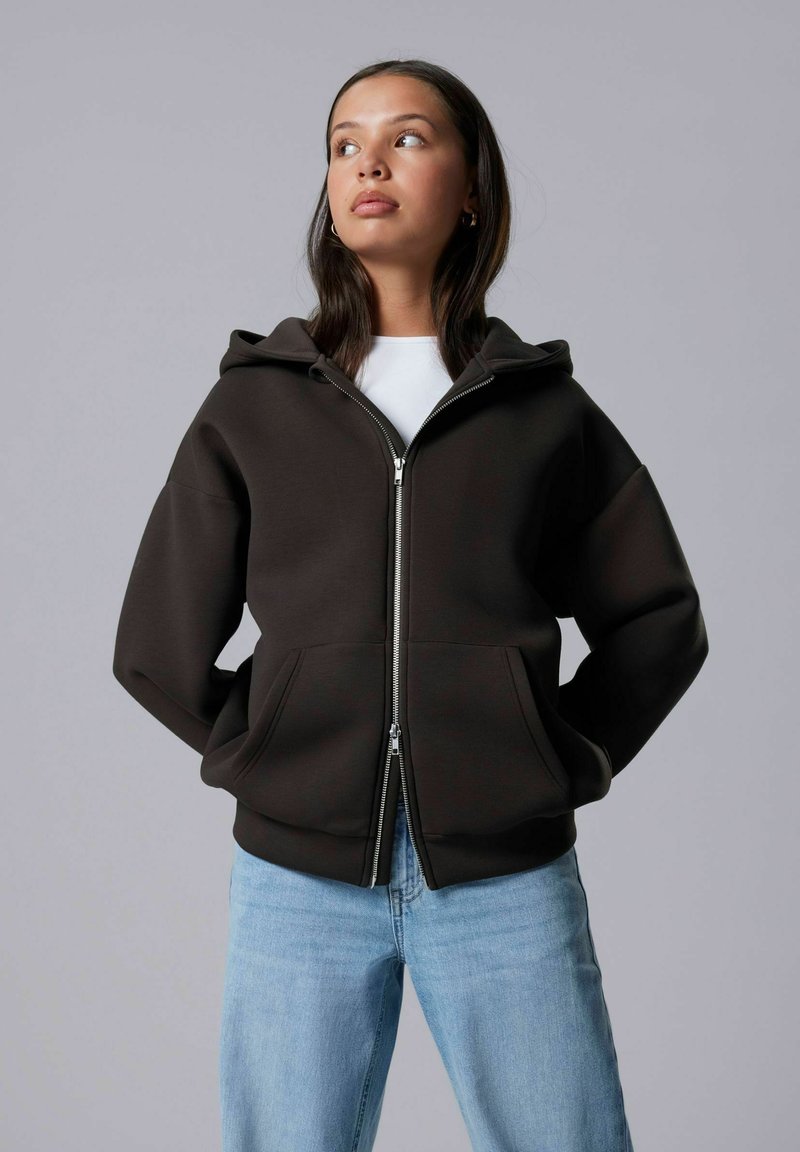 Felpa cappuccio marrone con zip frontale e due tasche, abbinata a jeans in denim azzurro chiaro. Tessuto liscio e vestibilità comoda.