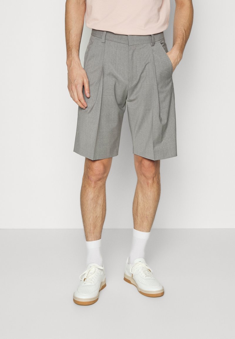 Homme portant un short plissé gris clair, des chaussettes blanches et des baskets blanches, debout avec une main dans la poche sur fond uni.
