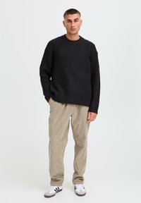 Czarny sweter prążkowany z okrągłym dekoltem, w zestawieniu z jasnymi, fakturowanymi spodniami z korduroju. Model nosi sportowe białe buty z czarnymi paskami.