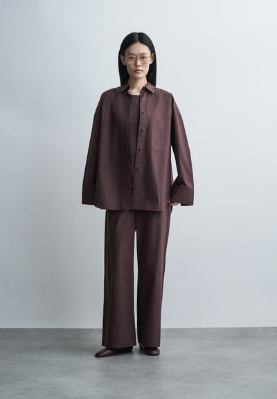 Femme aux longs cheveux noirs et lunettes, portant une chemise boutonnée marron foncé ample et un pantalon à jambes larges, debout sur un sol gris contre un mur clair.