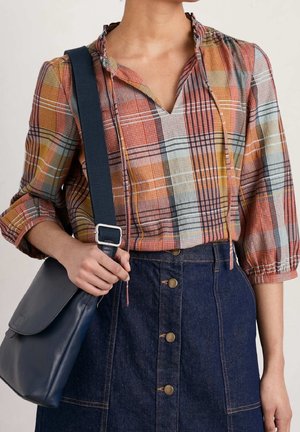 Persoon draagt een meerkleurige geruite blouse met opgerolde mouwen, een donkerblauwe spijkerrok met knopen aan de voorkant en houdt een navy schoudertas vast.