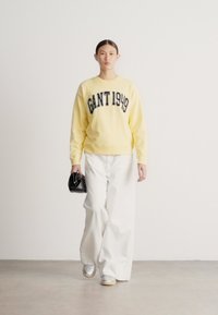 Sárga pulóver, rajta "GANT 1949" sötétkék betűkkel, laza fehér nadrággal és ezüst tornacipővel párosítva. A modell egy fekete kézitáskát tart.