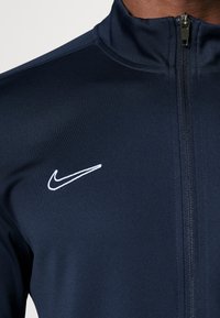 Marine blauwe ritsjack gemaakt van gestructureerde stof, met een verhoogde kraag en een witte geborduurde Nike swoosh op de linkerborst.