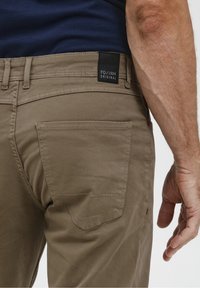 Jasnobrązowe spodnie o gładkiej fakturze. Posiadają tylną kieszeń naszywaną, czarną metkę i kontrastowe przeszycia w klasycznym stylu.