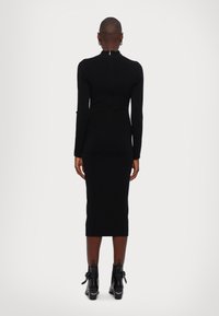 Personne avec les cheveux courts portant une robe midi noire ajustée à manches longues et des bottines noires, se tenant de dos contre un fond uni.
