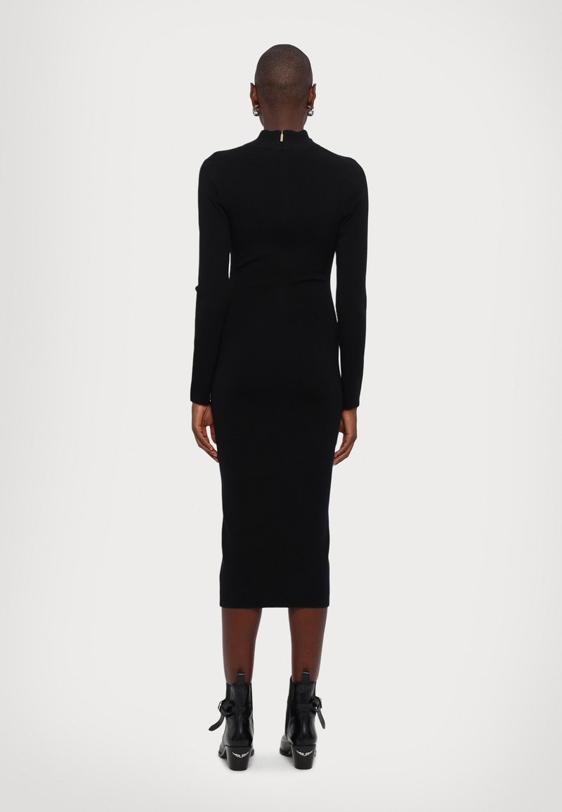 Persona con capelli corti che indossa un abito midi nero aderente a maniche lunghe e stivali neri alla caviglia, in piedi rivolto all'indietro su uno sfondo neutro.