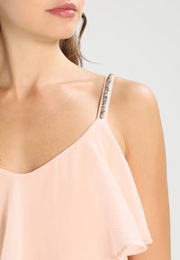 Světle růžový bezrukávový top s drapovaným designem a jemným zdobením po ramenním popruhu. Hladká, lehká tkanina.