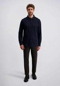 Man staat in een marineblauw overhemd met knopen, donkere bruine broek en zwarte nette schoenen tegen een effen lichte achtergrond.