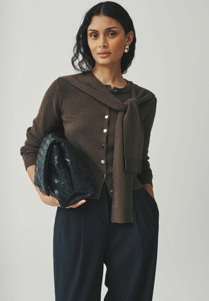 Brauner Cardigan mit Schalsdesign, ausgestattet mit goldenen Knöpfen und gerippten Bündchen, kombiniert mit einer strukturierten schwarzen geflochtenen Handtasche und marineblauen Hosen.