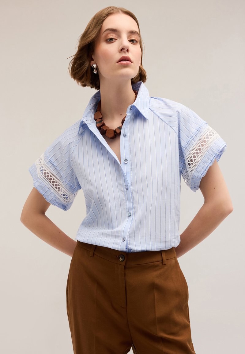 Jasnoniebieska koszula w paski z guzikami, z koronkowymi akcentami na rękawach, zestawiona z brązowymi spodniami. Posiada klasyczny kołnierzyk i luźny krój.