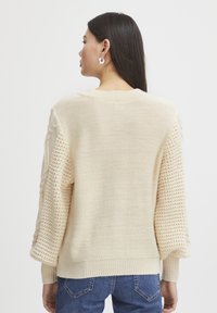 Pull en maille crème avec des manches texturées, présentant un ourlet côtelé et un col rond. Tissu doux avec une coupe décontractée.