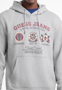 Felpa con cappuccio grigia con la scritta "GUESS JEANS", loghi circolari, tasca frontale e cordini. Il tessuto sembra morbido e casual.