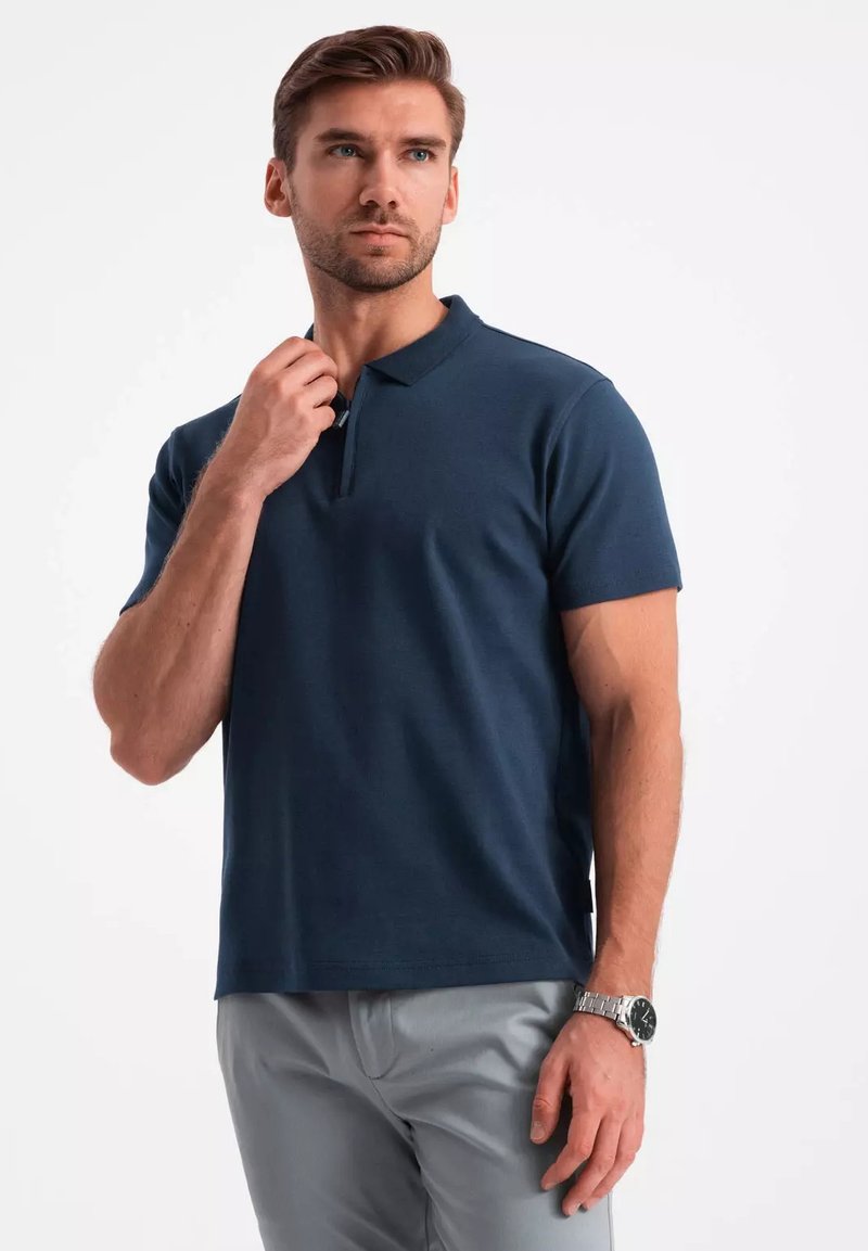 Granatowa koszulka polo wykonana z teksturowanej tkaniny, z zapięciem na trzy guziki i krótkimi rękawami. Połączona z jasnoszarymi spodniami i zegarkiem.