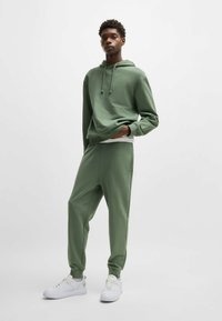Groene katoenen hoodie met een voorvak en koord, gecombineerd met bijpassende joggers. Model draagt witte sneakers en toont een ontspannen pasvorm.