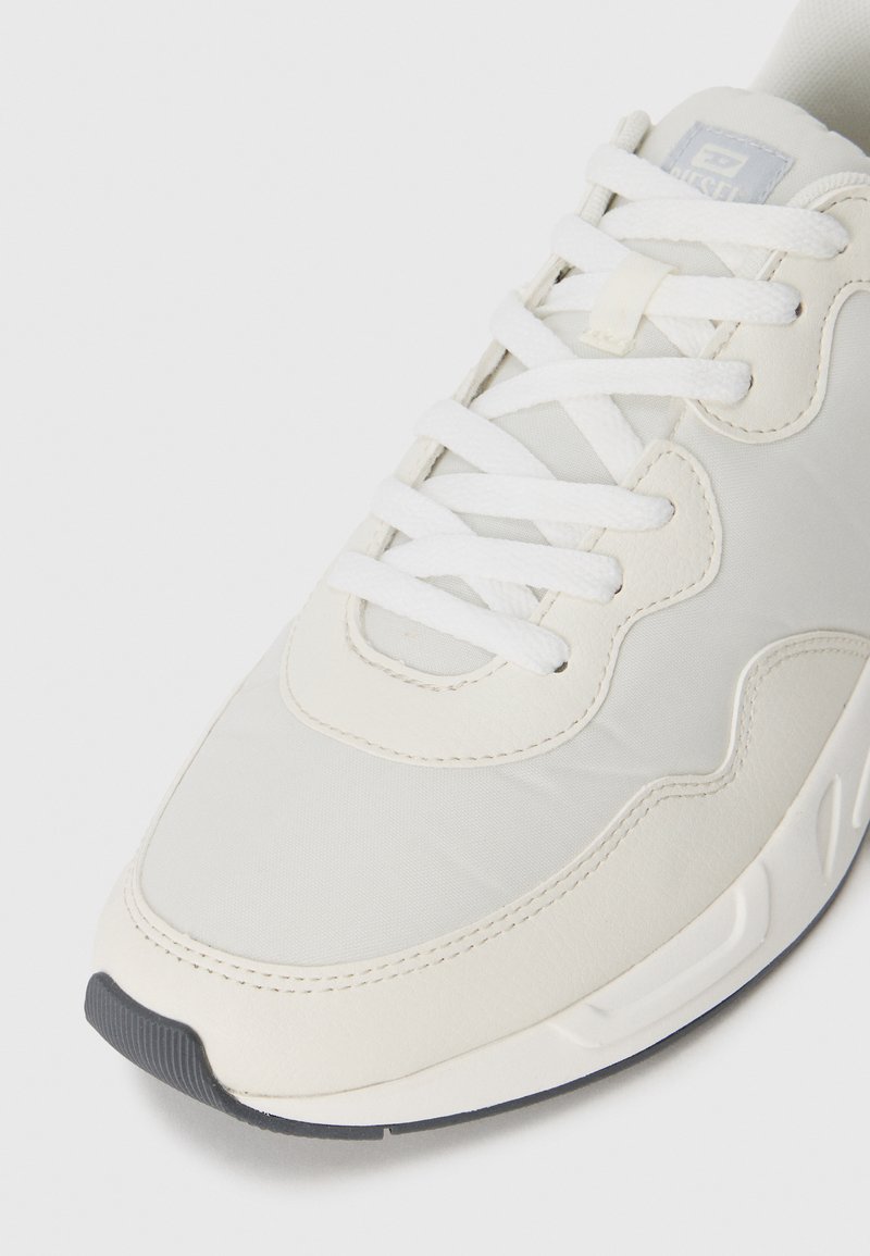 Zapatillas gris claro con un upper de tela texturizada, cordones blancos y una suela de goma. Presentan un parche con un logotipo sutil y acentos de diseño curvados.