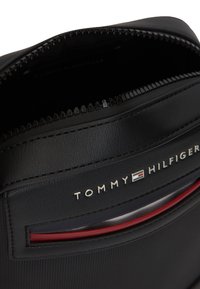 Μαύρη τσάντα Tommy Hilfiger με μερικώς ανοιχτό φερμουάρ, εμφανές λογότυπο και κόκκινη λεπτομέρεια στην μπροστινή τσέπη.